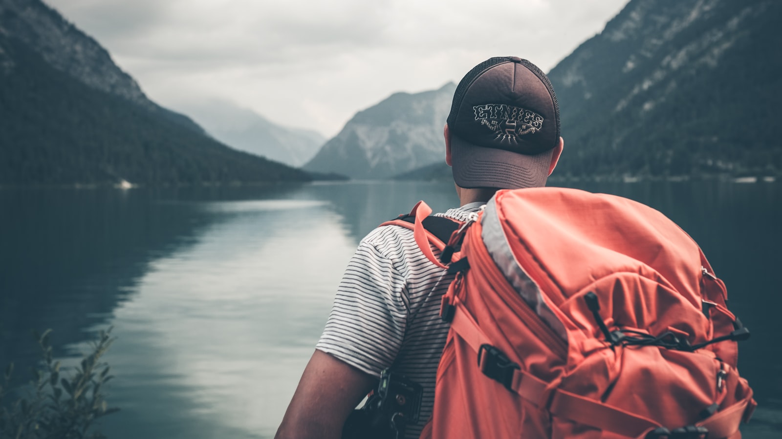 A traveller sitting on a wooden dock looking out over calm water with a small backpack beside them.